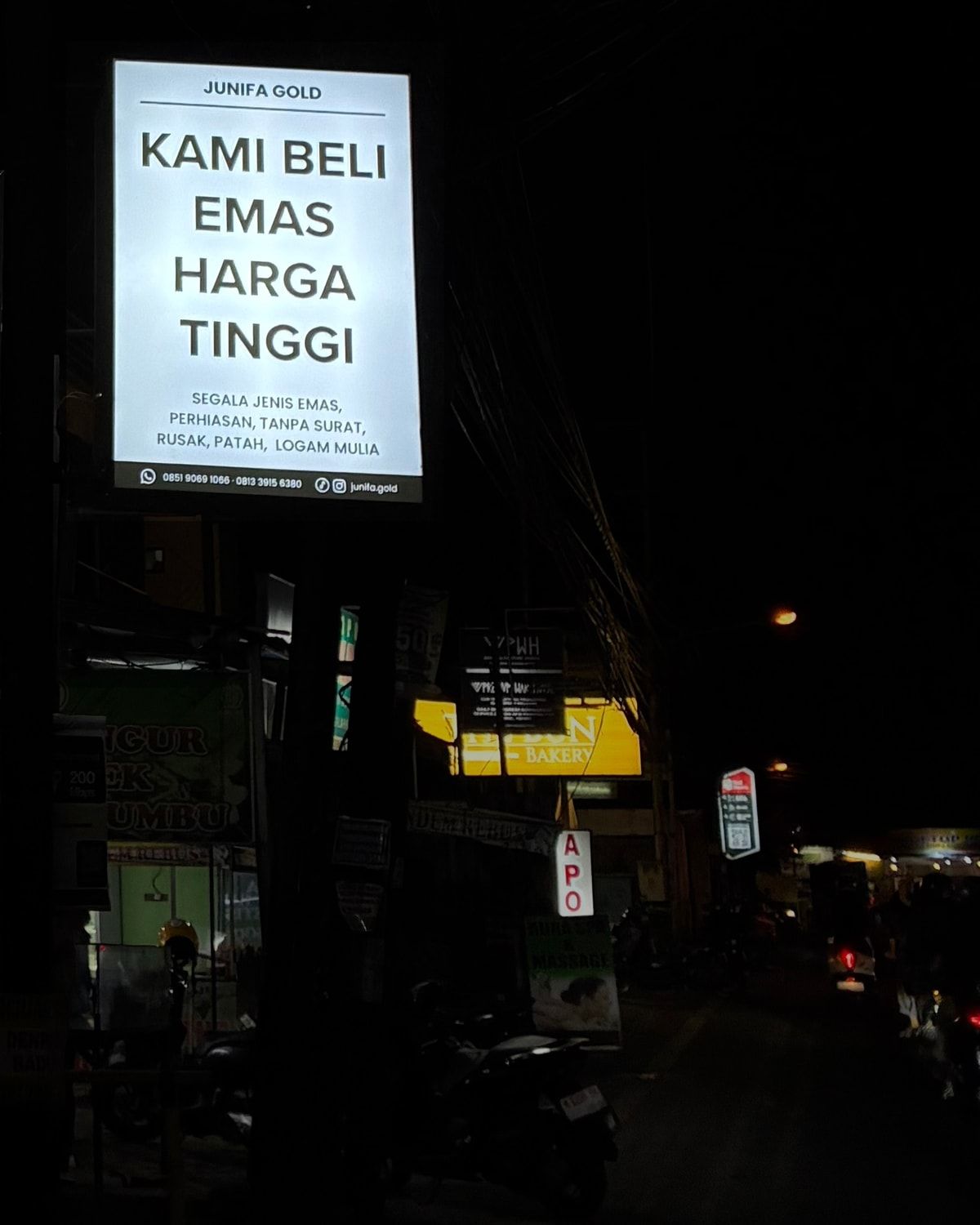 Papan nama Junifa Gold di pinggir jalan pada malam hari, bertuliskan Kami Beli Emas Harga Tinggi