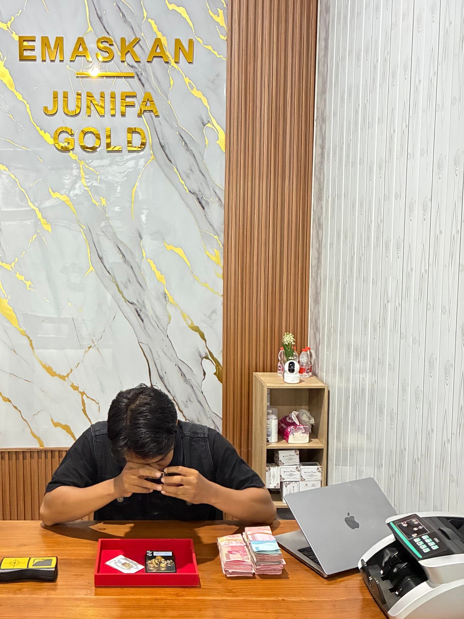 Tim Junifa Gold memeriksa perhiasan emas dengan kaca pembesar di meja transaksi, dilihat dari kursi penjual, dengan papan nama EMASKAN dan Junifa Gold di belakangnya
