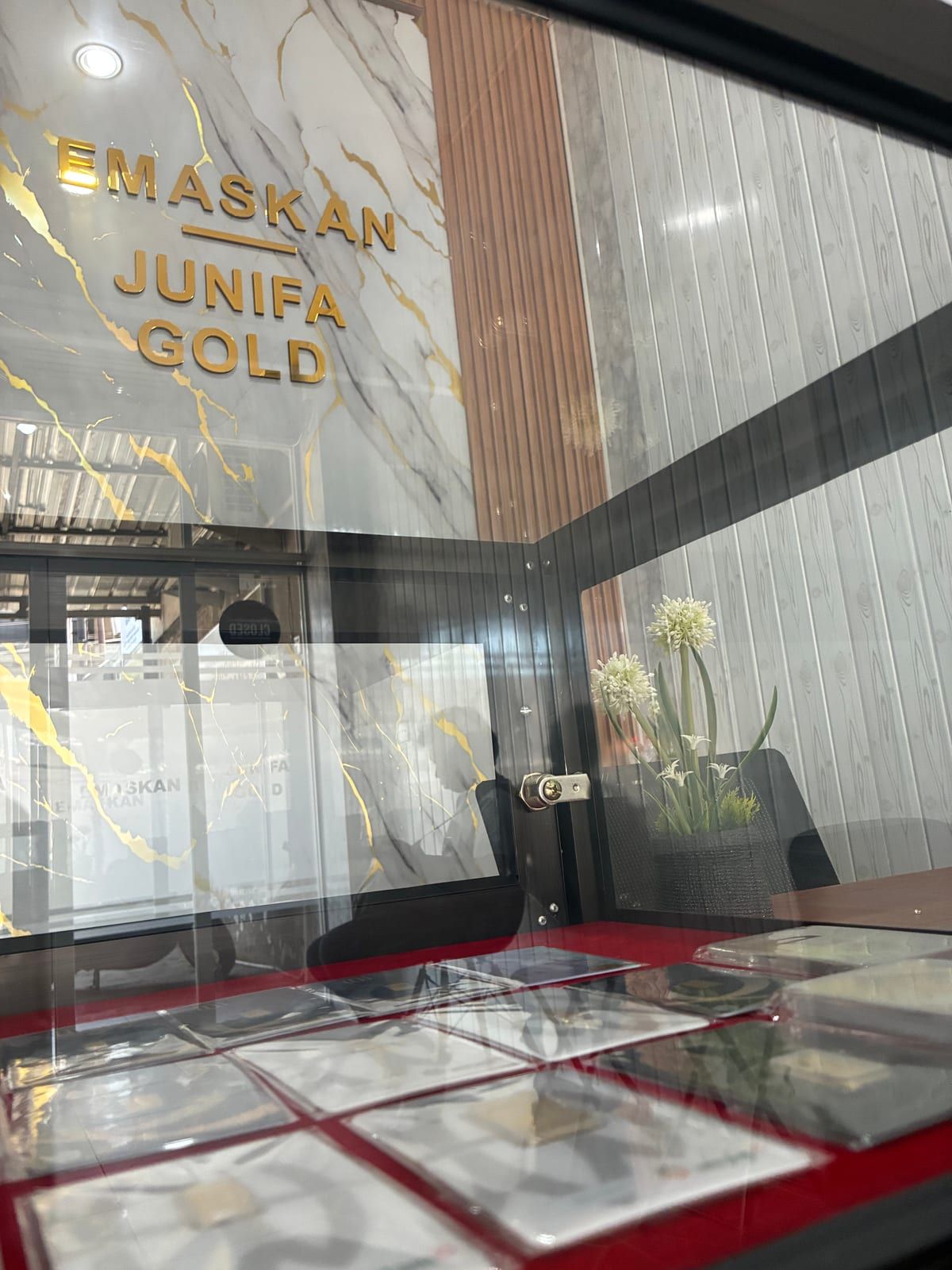 Interior toko Junifa Gold di Denpasar: papan nama EMASKAN dan Junifa Gold berhuruf emas di dinding marmer, dilihat dari balik etalase kaca